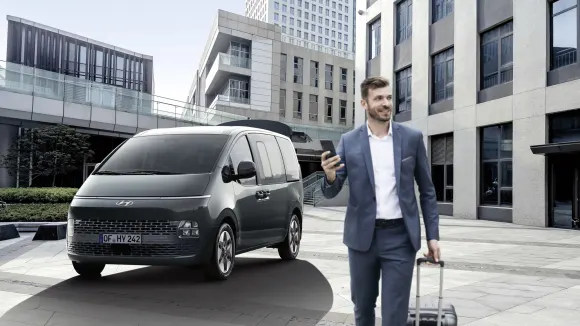 Ein Mann in Business-Anzug und Smartphone in der Hand vor seinem geparkten Hyundai STARIA.