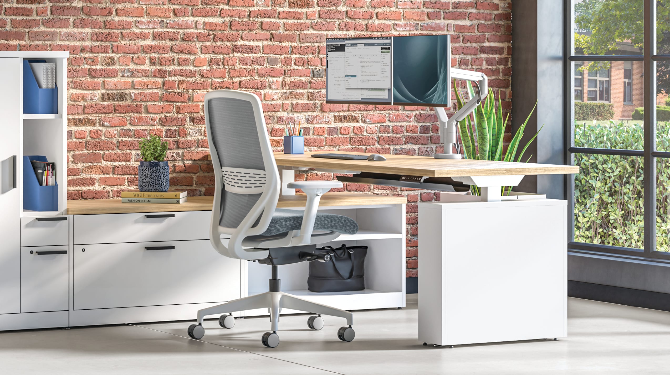 Open office workspace with white desks and ergonomic chairs, modular storage cabinets, computer monitors, and exposed brick wall with large windows.