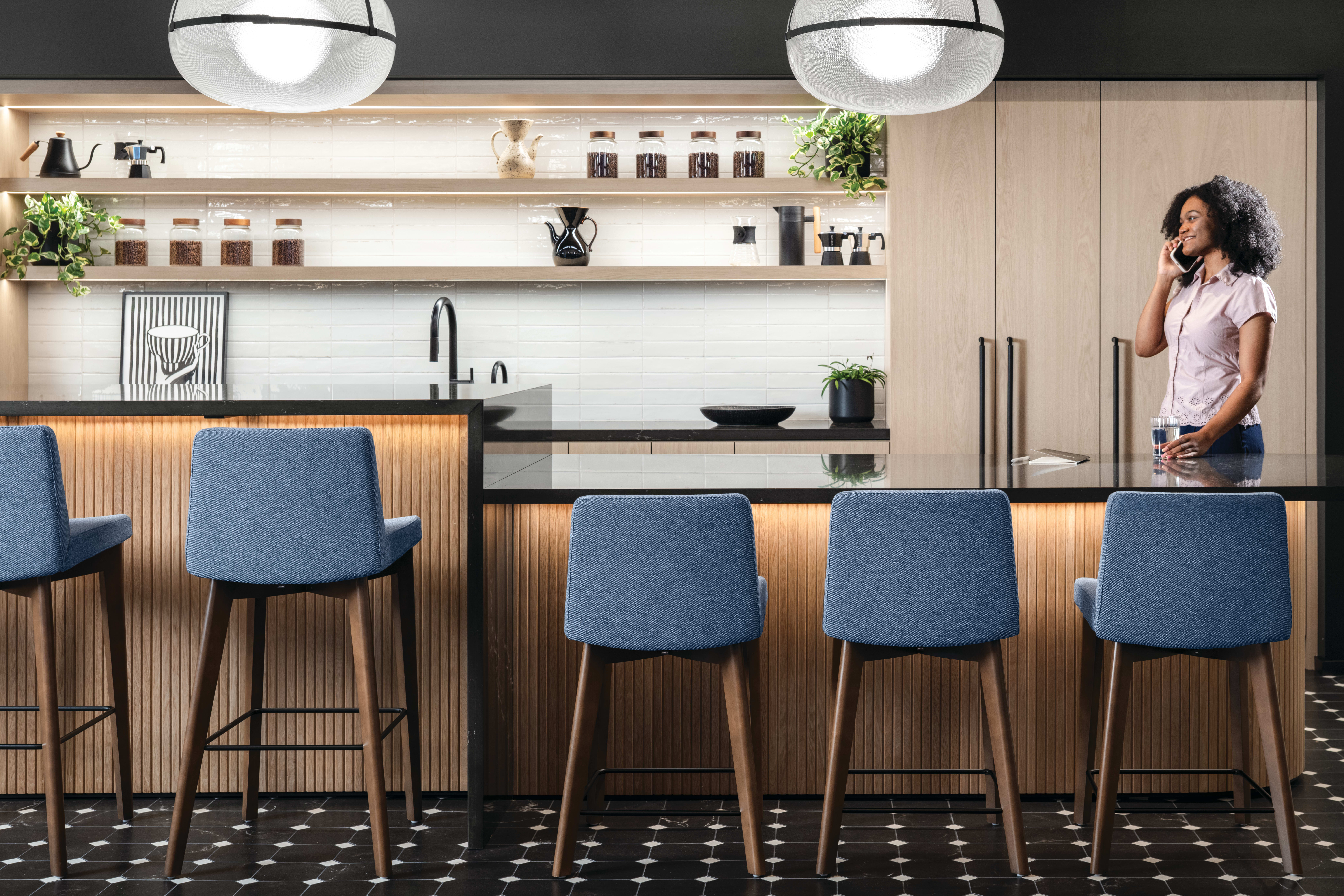 Cafe-style bar area with wood slat-front counter, black stone countertop, upholstered bar stools with wood legs, open shelving, and tall storage cabinets.