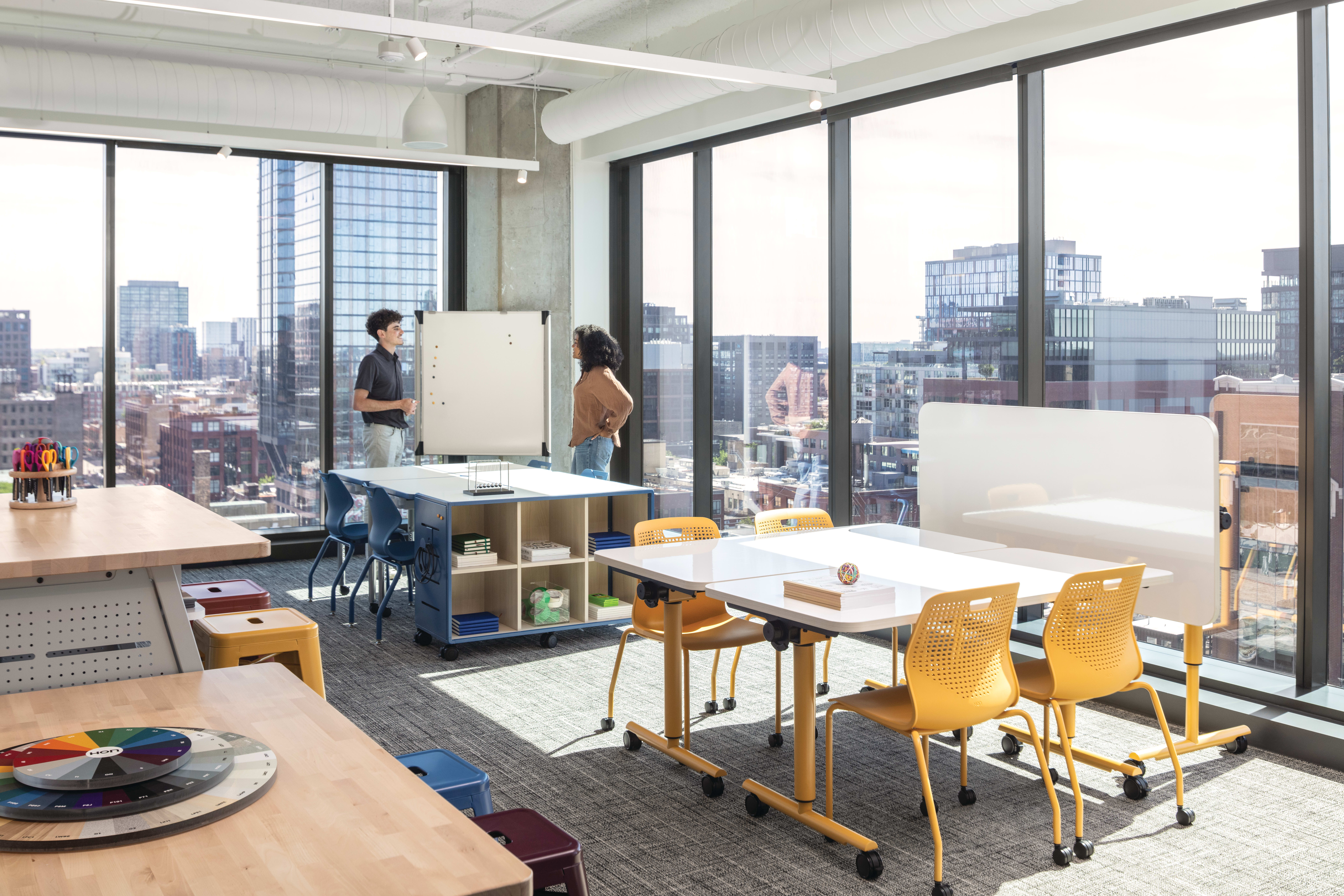 Flexible workspace with mobile tables, laminate work surfaces, caster-based storage unit, and stackable plastic chairs with perforated backs.