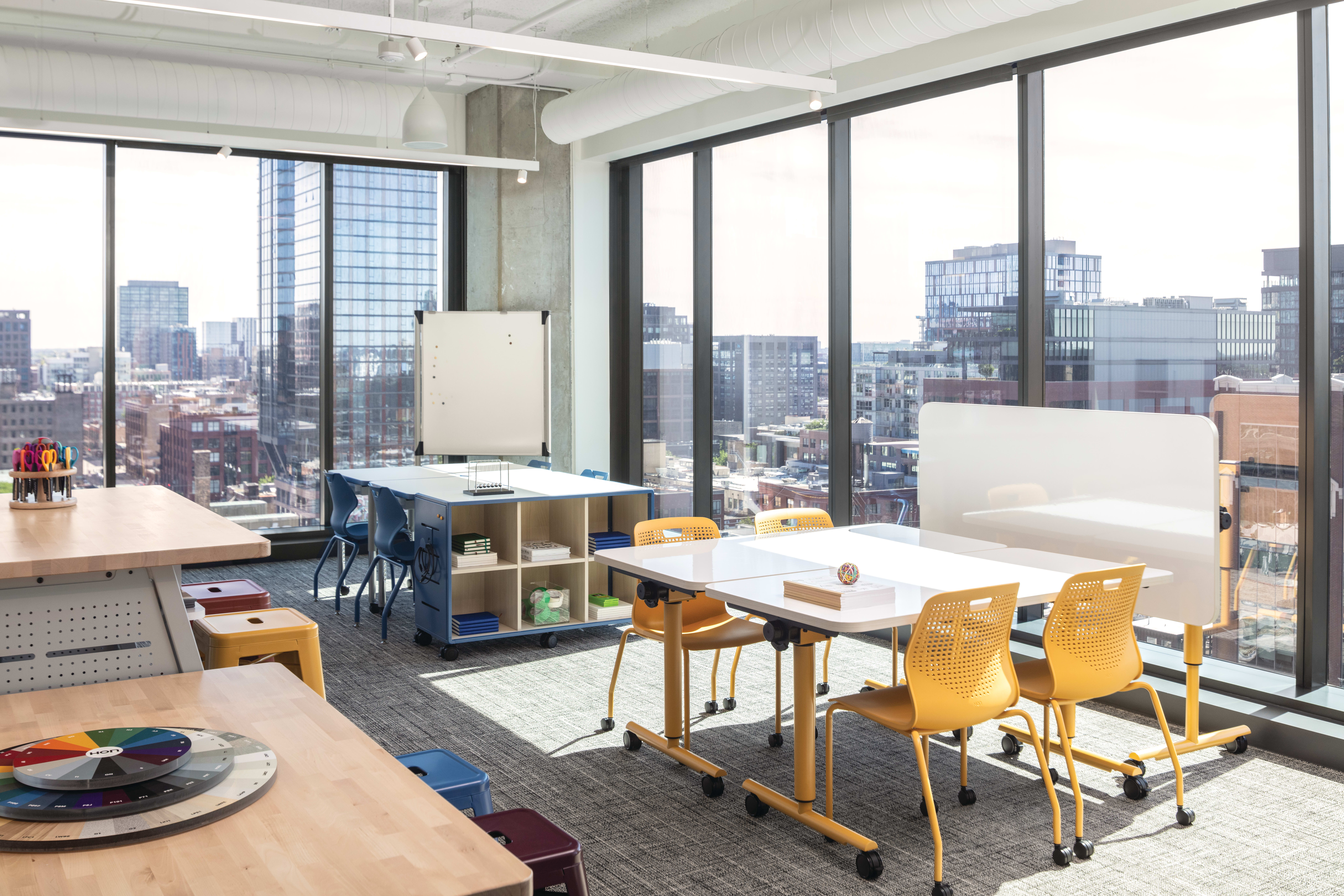 Flexible workspace with mobile tables, laminate work surfaces, caster-based storage unit, and stackable plastic chairs with perforated backs.