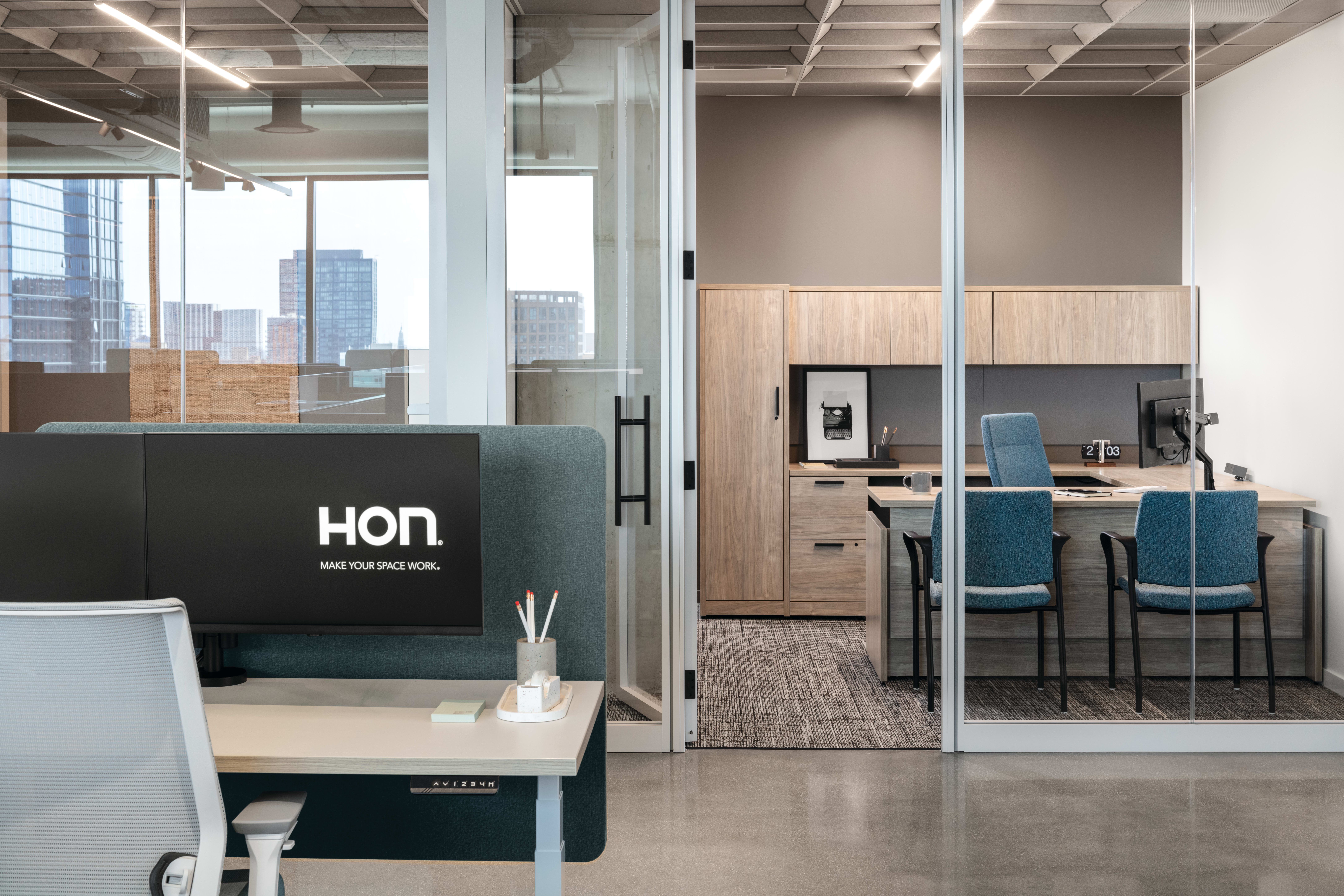 Open workstation with height-adjustable desk, privacy panel, task chair, and monitor, paired with glass-enclosed office featuring L‑shaped work surface, storage cabinets, and guest seating.