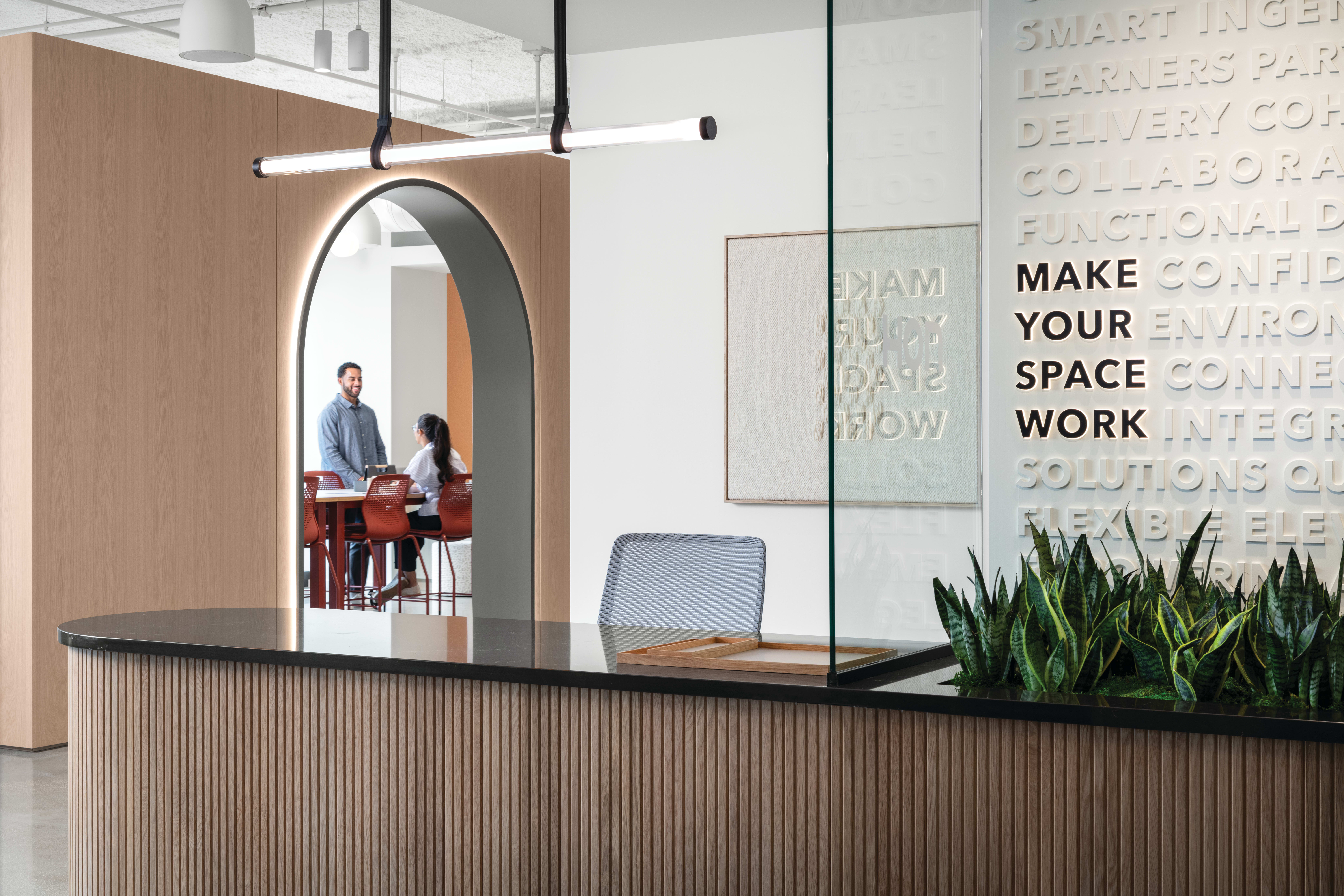 Reception counter with wood slat detailing and solid surface top, paired with task seating, integrated planter, pendant lighting, glass partition, and arched wall opening leading to collaborative seating area.