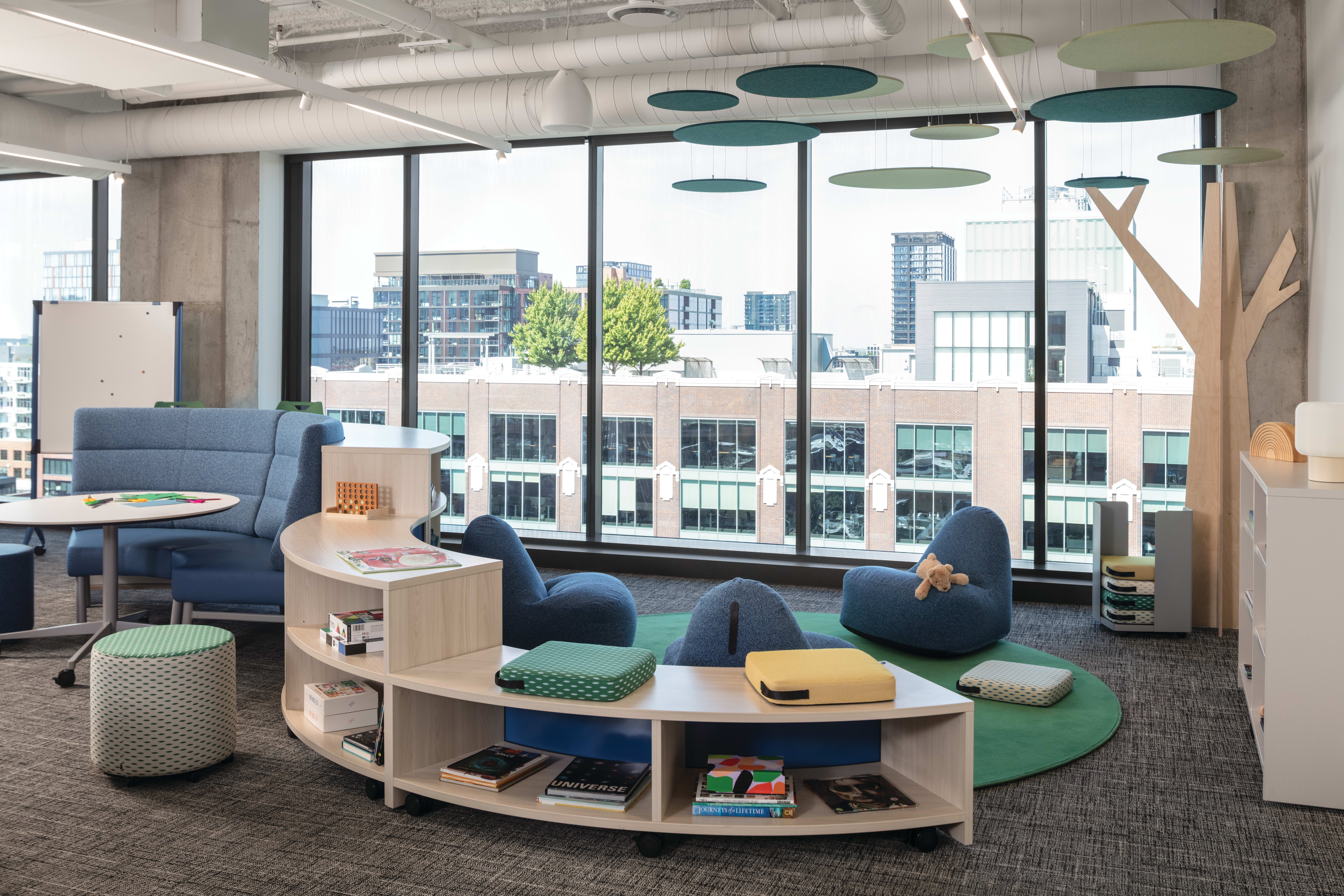 Flexible lounge area with upholstered seating, ottomans, low tables, open shelving, and acoustic ceiling panels near large windows.