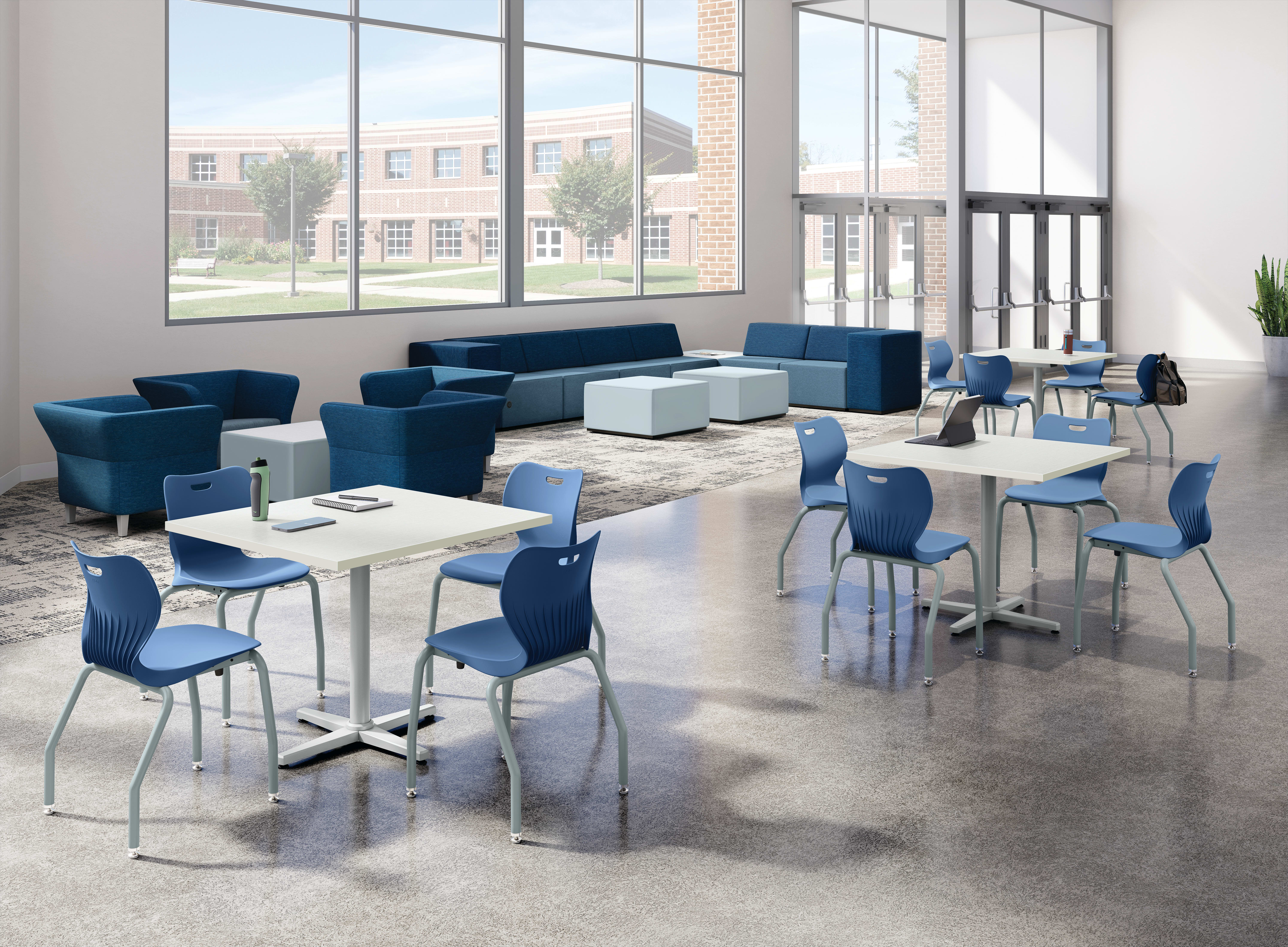 Bright student commons area with collaborative tables, blue classroom chairs, lounge seating, floor-to-ceiling windows, polished concrete flooring, and open gathering space in a modern educational environment.
