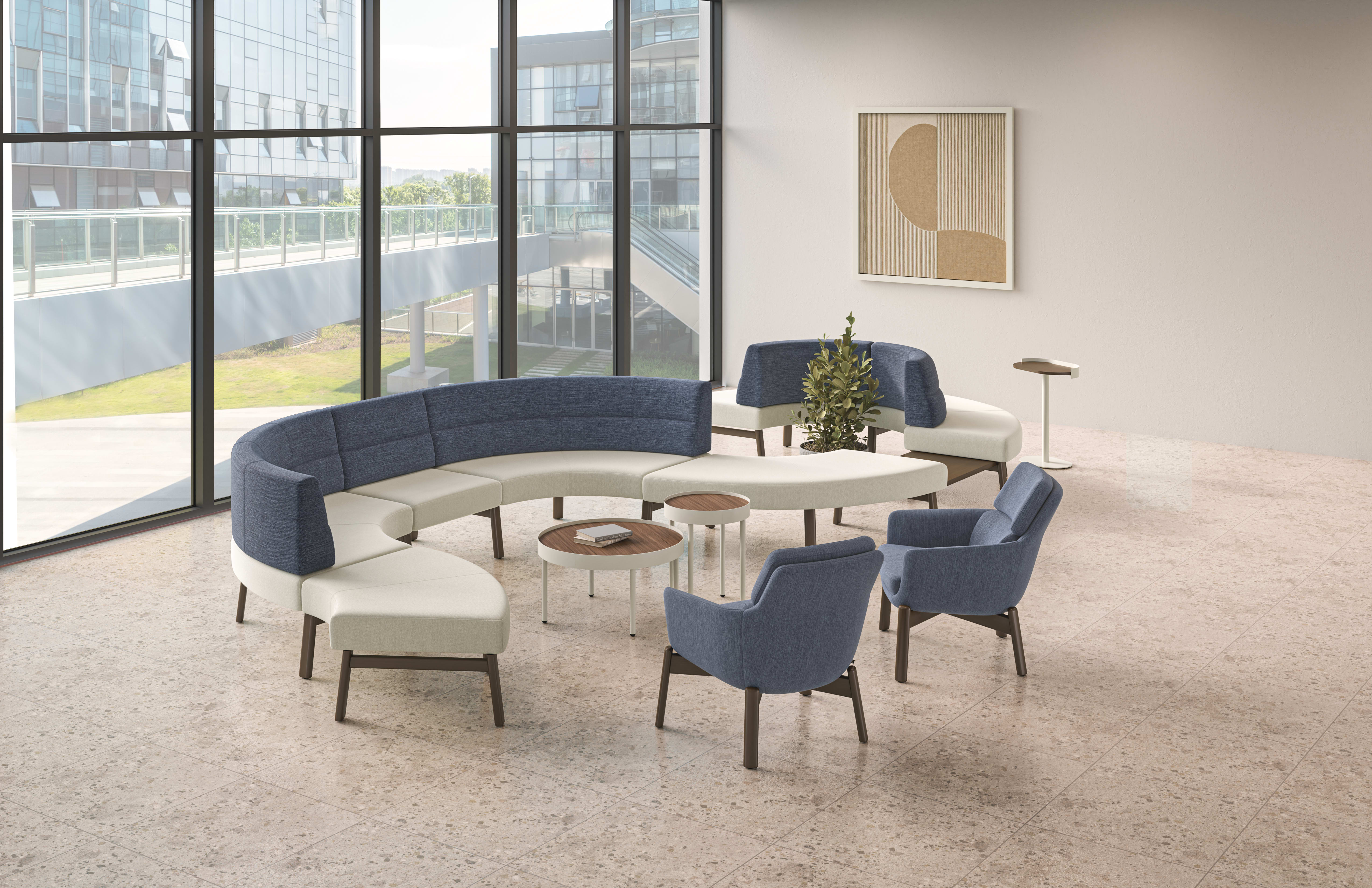 Modern lounge area with curved blue and white modular seating, blue lounge chairs, round wood accent tables, and floor-to-ceiling windows in an open collaborative workspace.