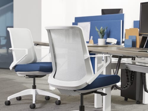 The image shows a modern office space with white and blue office chairs arranged around a table, with shelves and other office equipment visible in the background.
