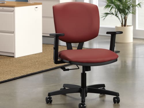 A red office chair with adjustable armrests sits on a tiled floor, with a filing cabinet and a potted plant visible in the background.