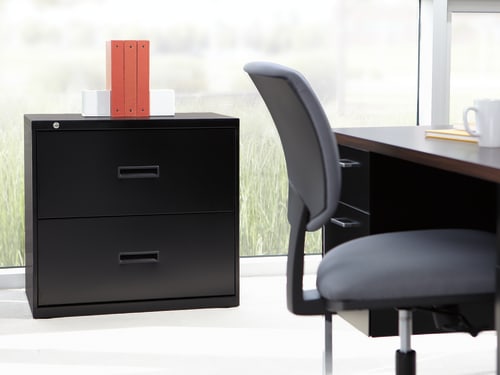 A black filing cabinet stands in the foreground, with a red folder on top, next to a swivel chair in front of a large window overlooking a grassy outdoor scene.