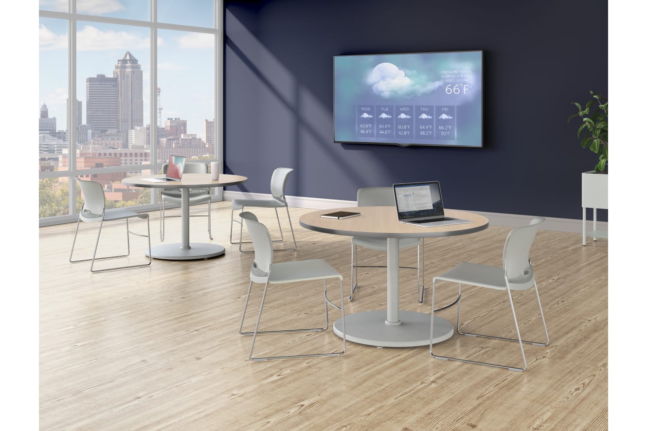 A bright lounge area with round tables, light-colored chairs, a wall-mounted display showing weather information, and large windows overlooking a city skyline.