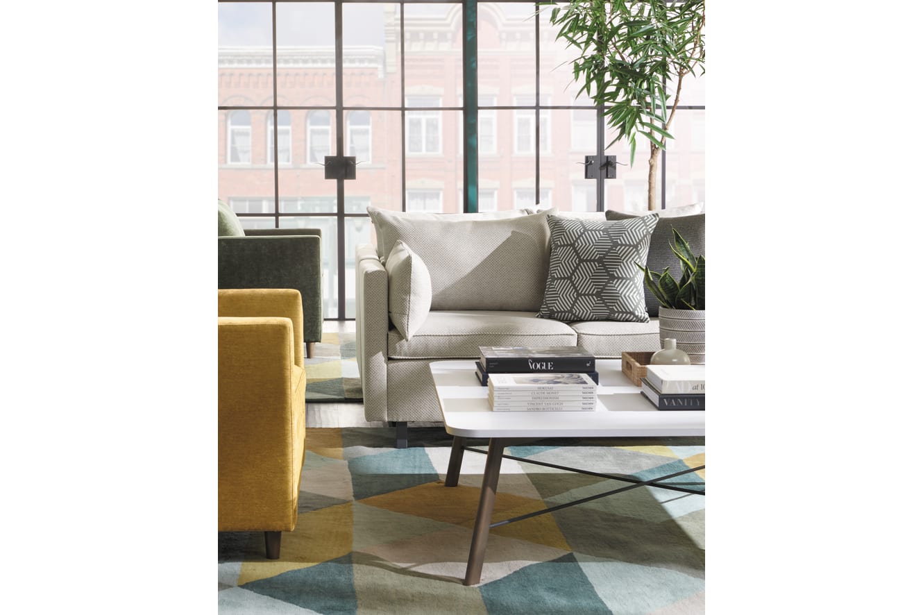 A cozy and modern living room with a beige sofa, patterned throw pillows, a yellow armchair, and a glass coffee table on a colorful area rug, set against a backdrop of large windows overlooking a city skyline.