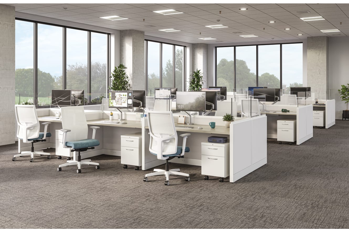 Open office layout with white workstations, ergonomic chairs, desktop monitors, and large windows overlooking greenery.