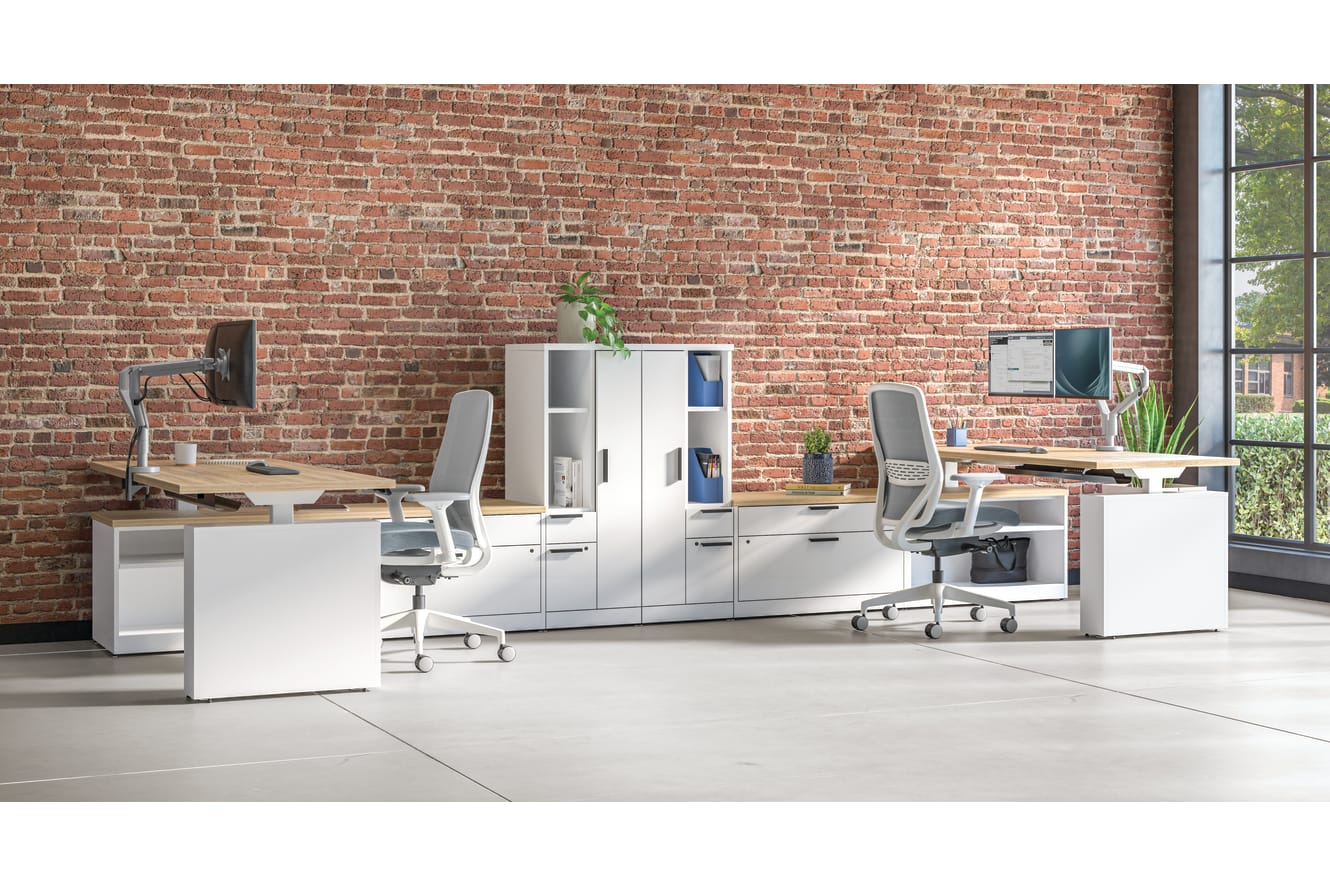 Open office workspace with white desks and ergonomic chairs, modular storage cabinets, computer monitors, and exposed brick wall with large windows.