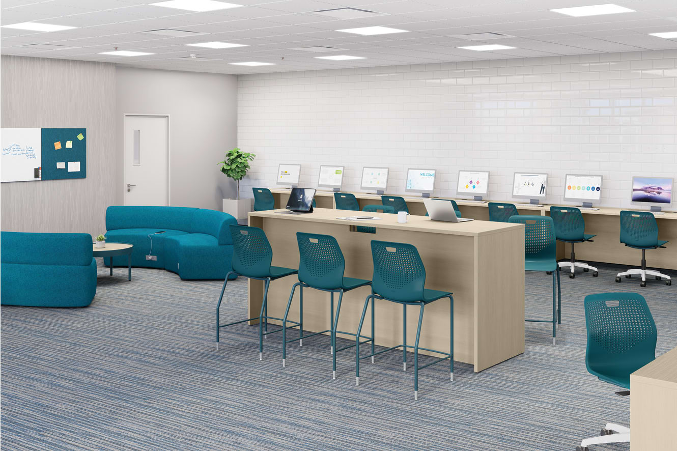Modern computer lab with long worktables, desktop monitors, laptops, teal task chairs, soft seating area, white brick accent wall, carpeted flooring, and bright overhead lighting.