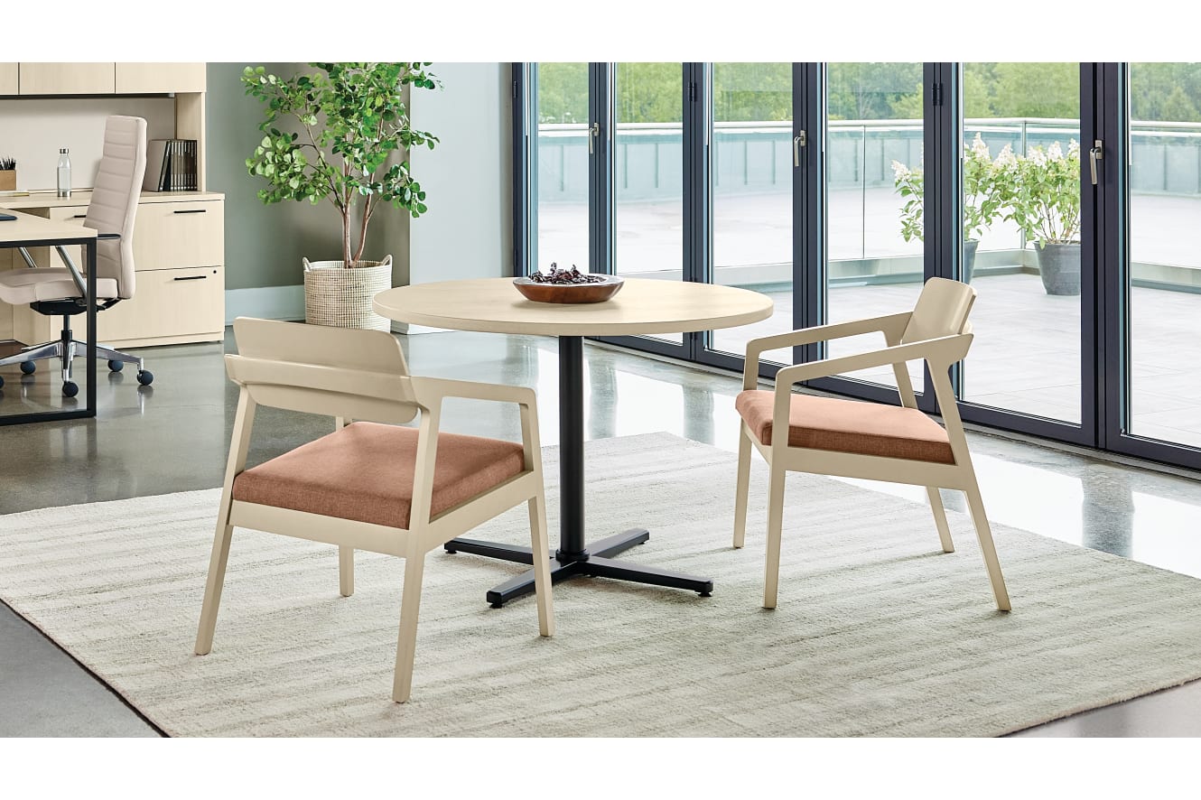 A modern, minimalist dining area with a round wooden table, beige chairs, and a large window overlooking a lush, green outdoor space.