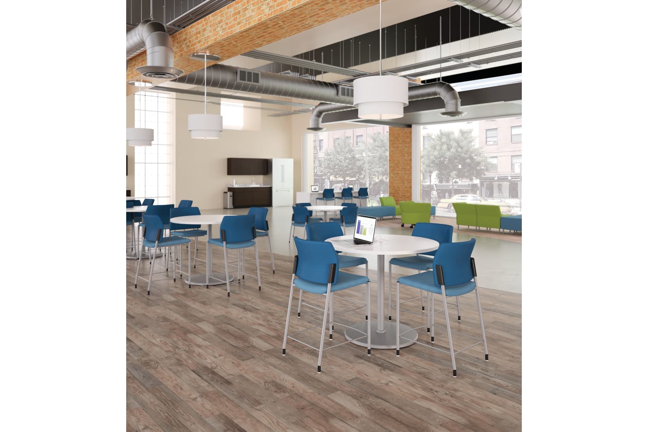 A modern and open office space with a mix of blue and green chairs, round tables, and exposed ceiling ducts, creating a bright and collaborative work environment.
