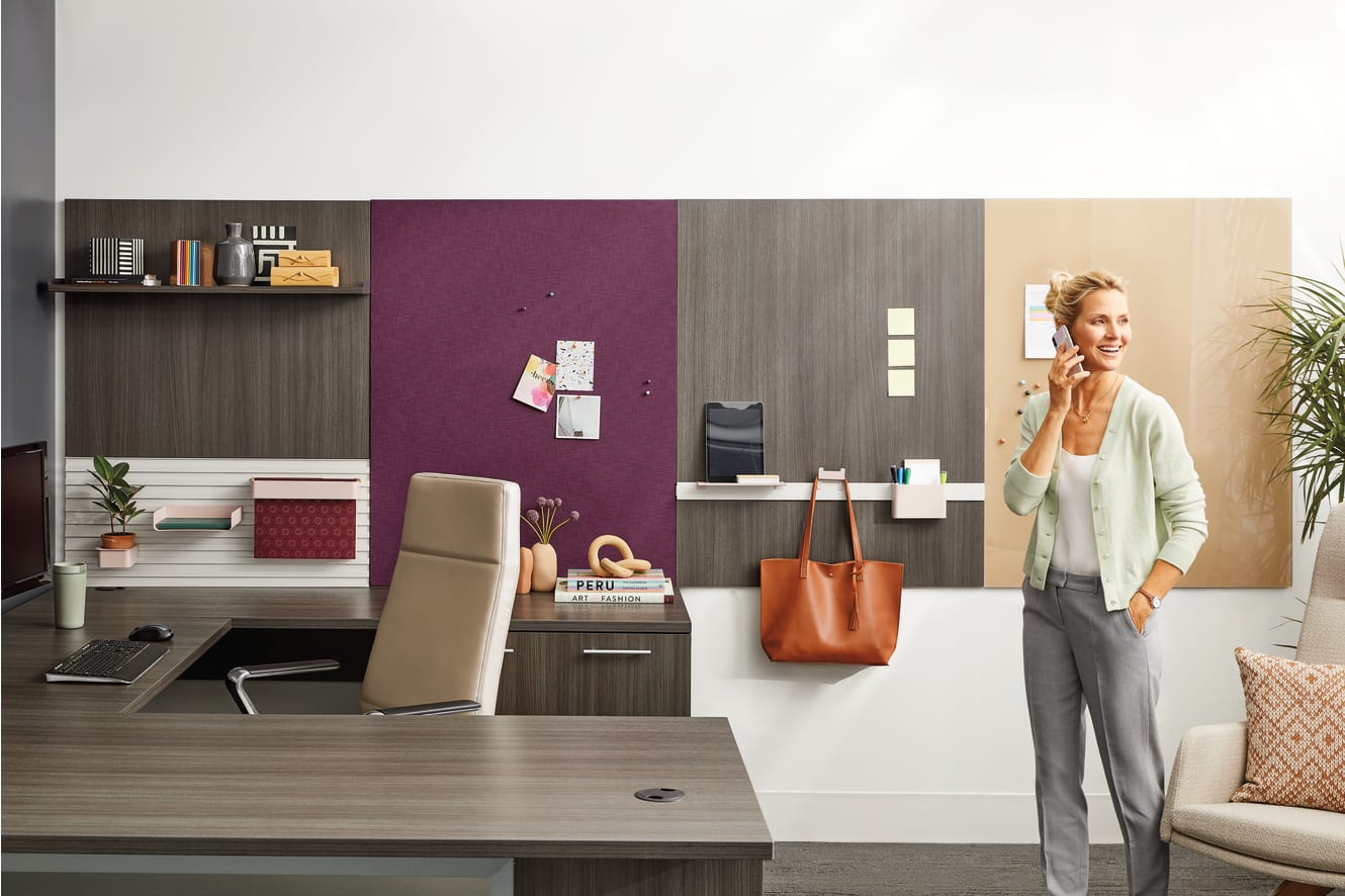 A woman stands in a modern, well-furnished home office, surrounded by shelves, a desk, and various decor items.