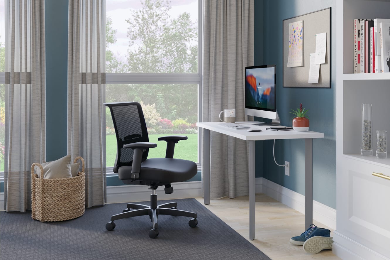 A modern home office with a white desk, a black ergonomic office chair, and various decorative elements such as artwork, plants, and a woven basket, set against a backdrop of large windows overlooking a lush outdoor scene.