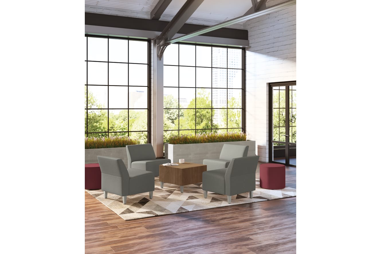 A modern and spacious living room with large windows overlooking a lush outdoor landscape, featuring comfortable gray armchairs, a wooden coffee table, and a patterned area rug on the hardwood floor.