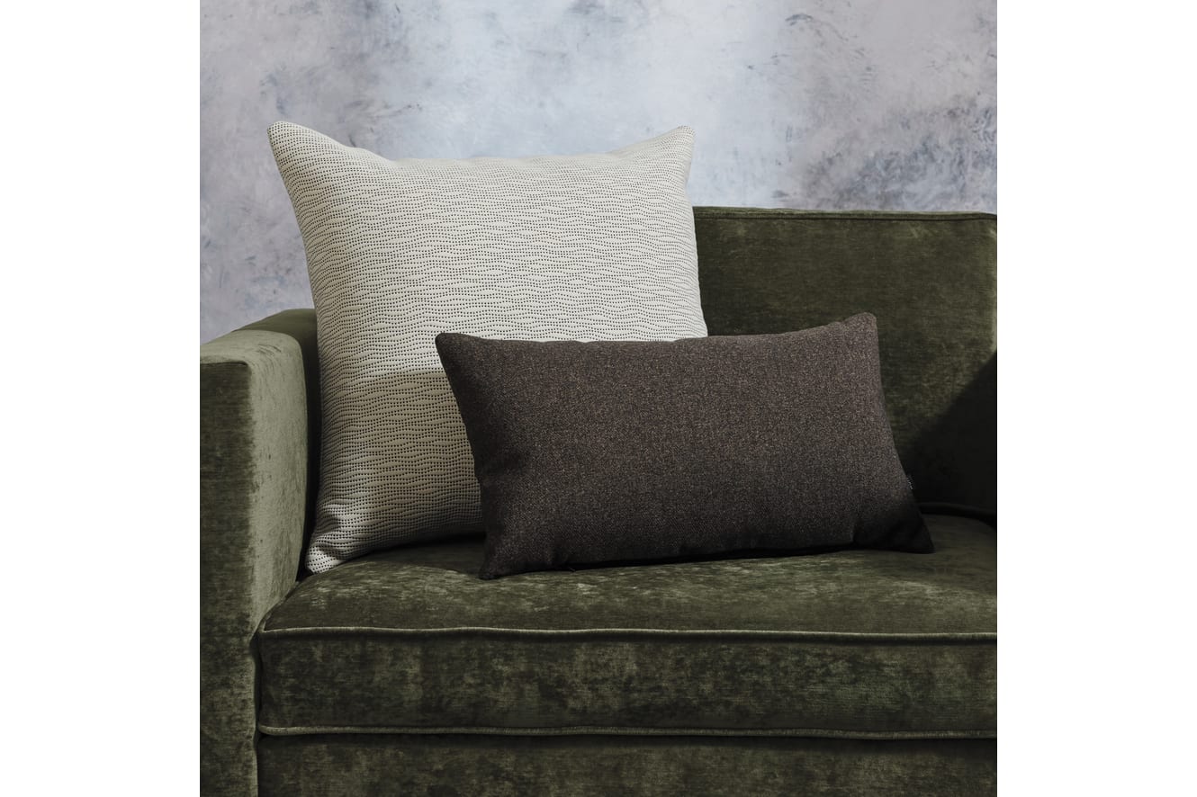 The image shows a green velvet sofa with two decorative pillows, one in a light beige pattern and the other in a dark brown solid color, against a plain gray background.