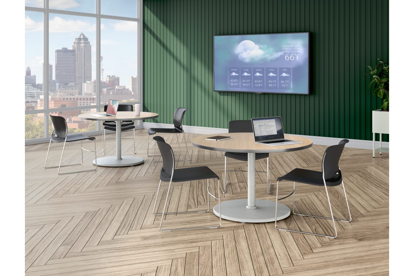 A modern lounge area with round tables, black chairs, a large wall-mounted display showing weather information, and floor-to-ceiling windows overlooking a city skyline.