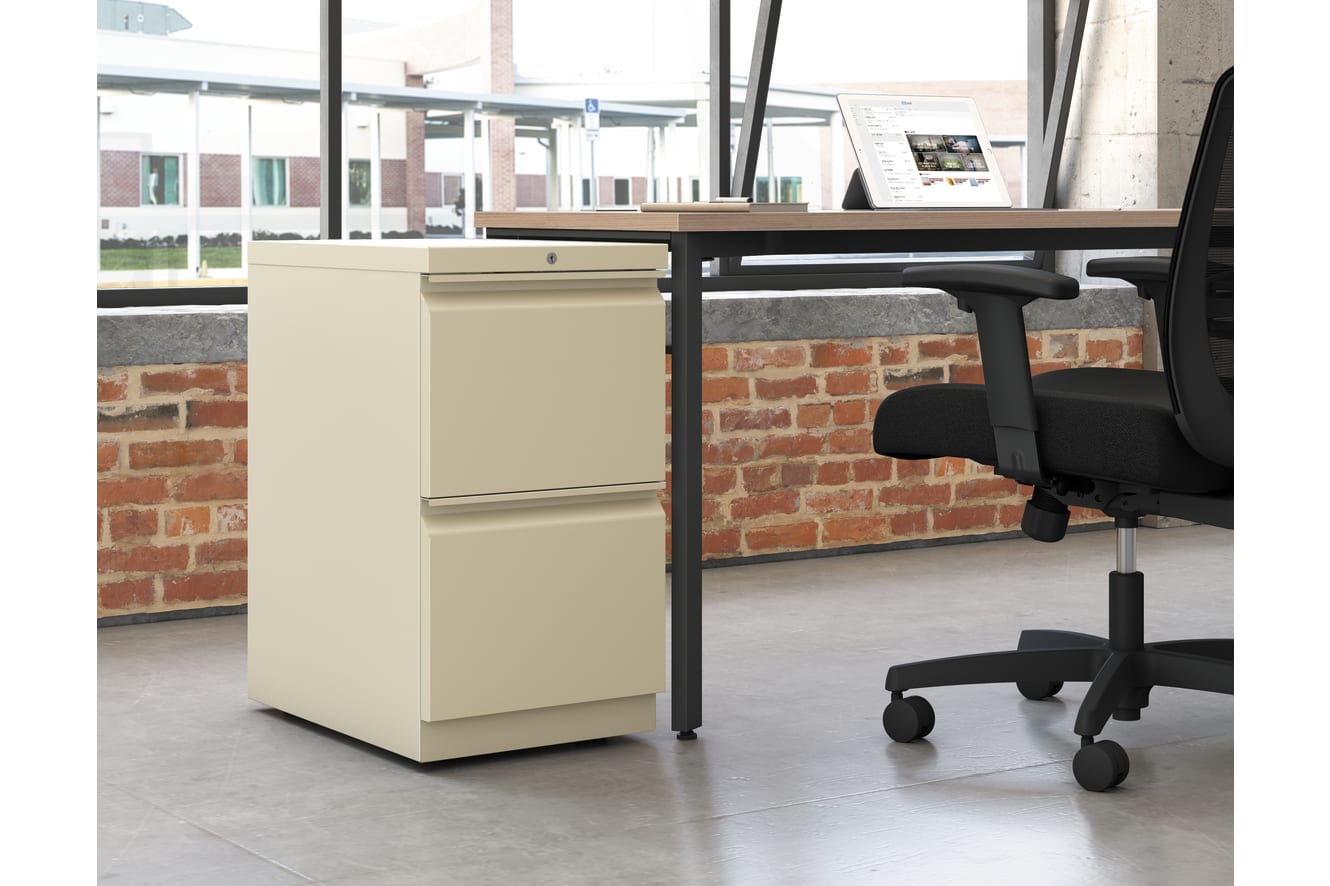 A modern office workspace with a wooden desk, a beige filing cabinet, and a black office chair set against a backdrop of exposed brick walls and large windows.