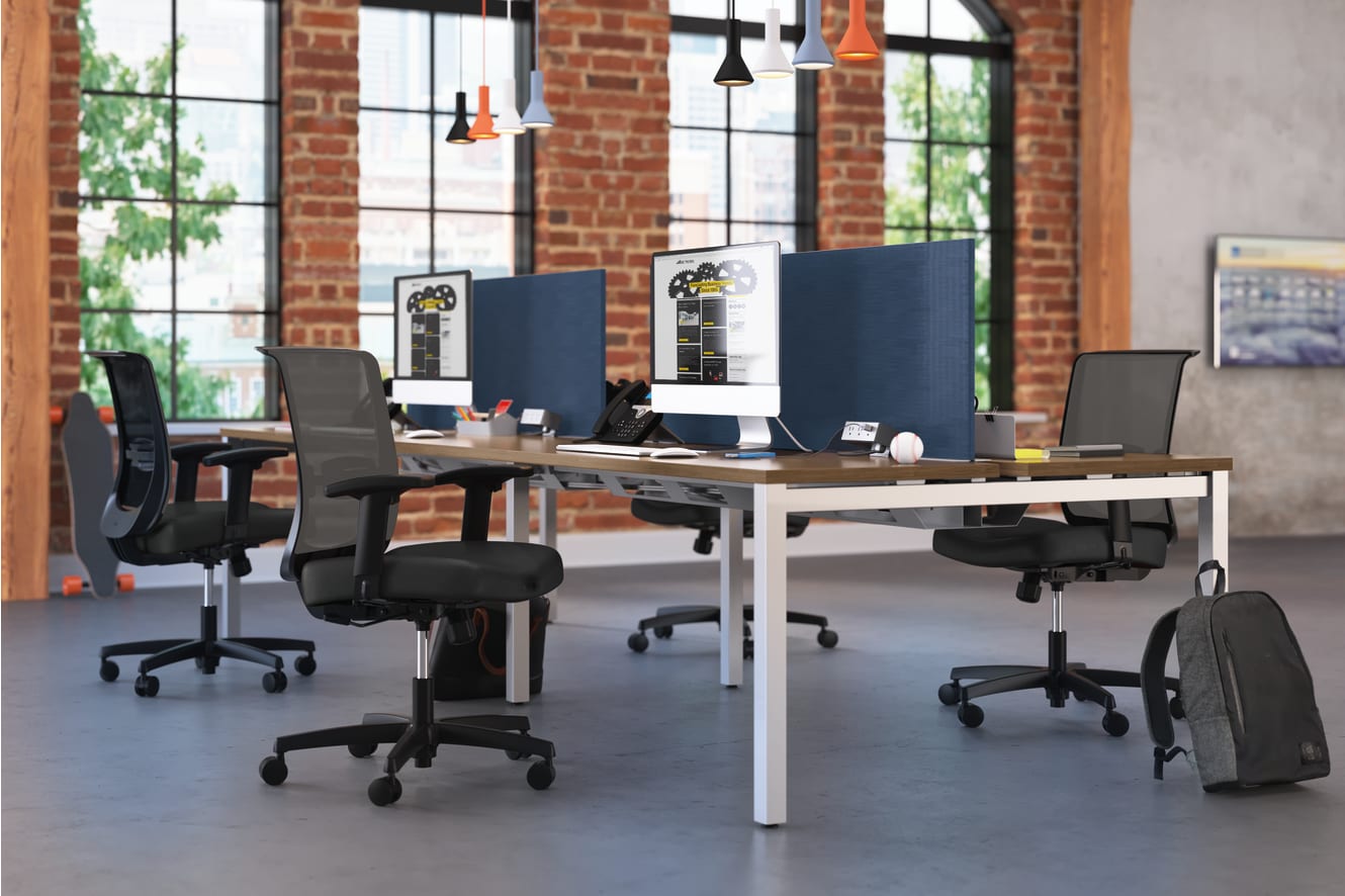 A modern, open-plan office space with brick walls, large windows, and a long wooden desk with multiple computer workstations and office chairs.