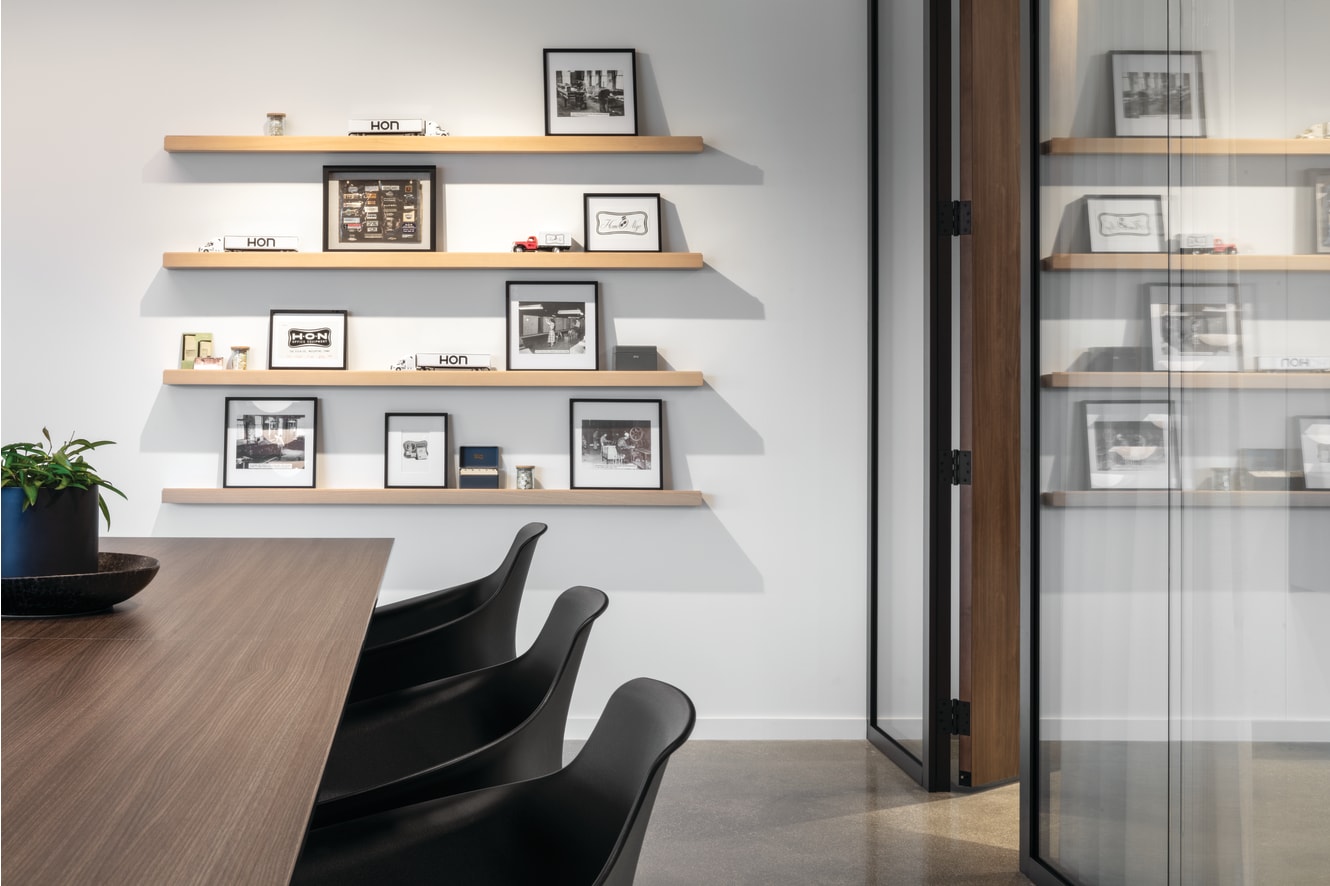 Copilot said: Conference space with wood meeting table, molded guest chairs, glass partition wall, and floating wood shelves displaying framed prints and accessories.
