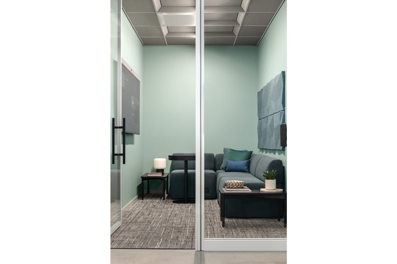 Copilot said: Glass-enclosed focus room with upholstered lounge seating, accent pillows, side tables, integrated media display, acoustic wall panels, and soft lighting.