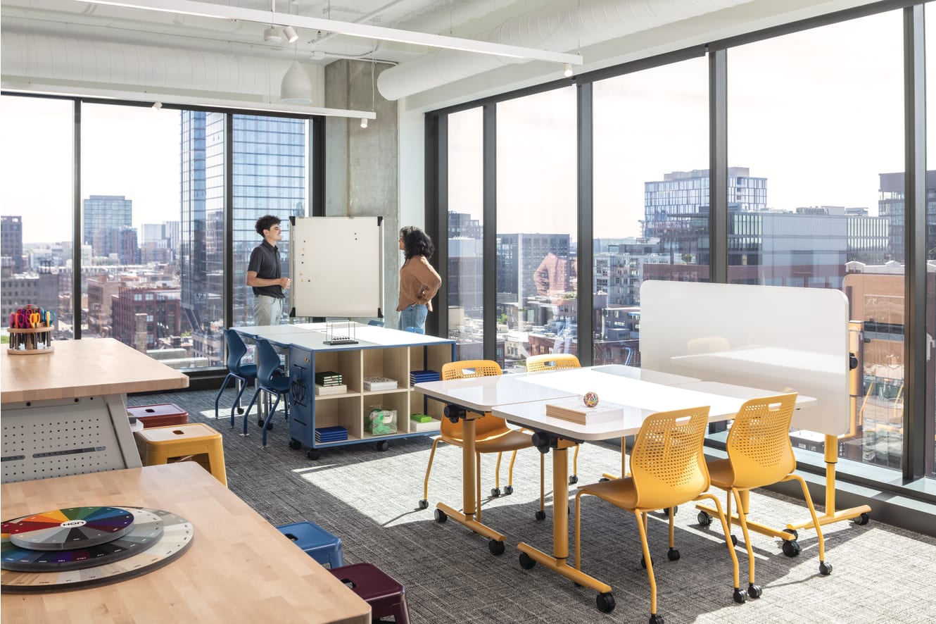 Flexible workspace with mobile tables, laminate work surfaces, caster-based storage unit, and stackable plastic chairs with perforated backs.