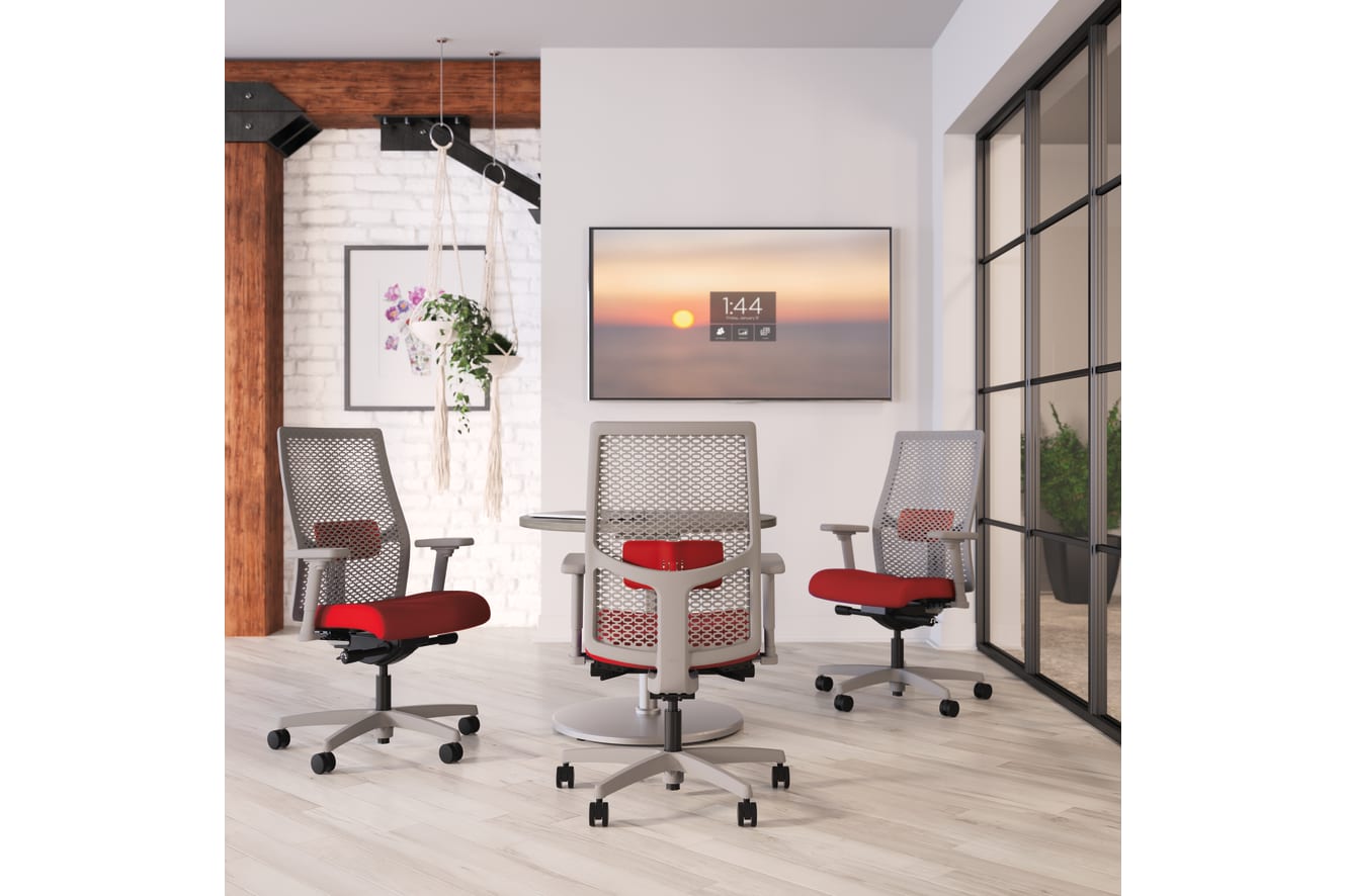 The image depicts a modern office space with white walls, wooden accents, and large windows overlooking a scenic sunset. In the foreground, there are several red and gray office chairs arranged around a table.
