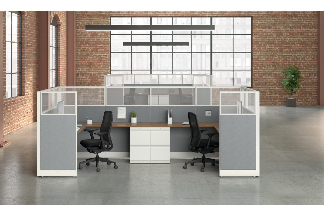 A modern, minimalist office space with white and gray cubicles, chairs, and a potted plant against a brick wall background.