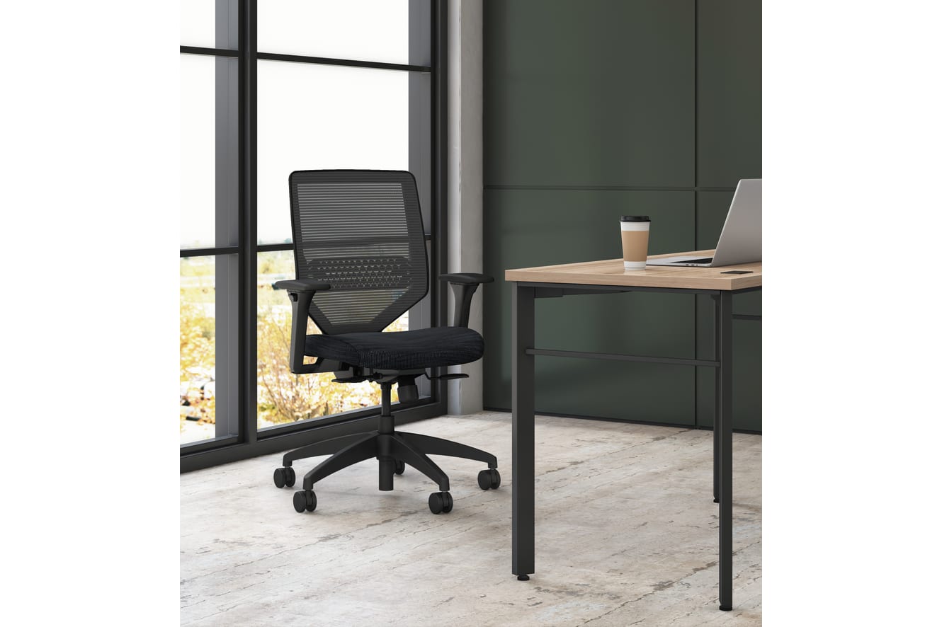 A modern, minimalist office space with a black mesh office chair and a wooden desk with a laptop and coffee cup, set against a large window overlooking a natural outdoor scene.