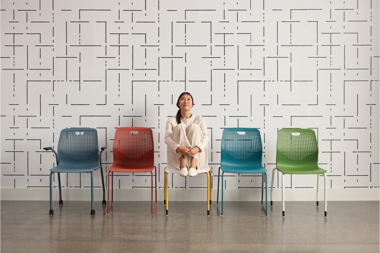 A woman in a white outfit sits on a chair in front of a patterned wall, surrounded by colorful chairs.