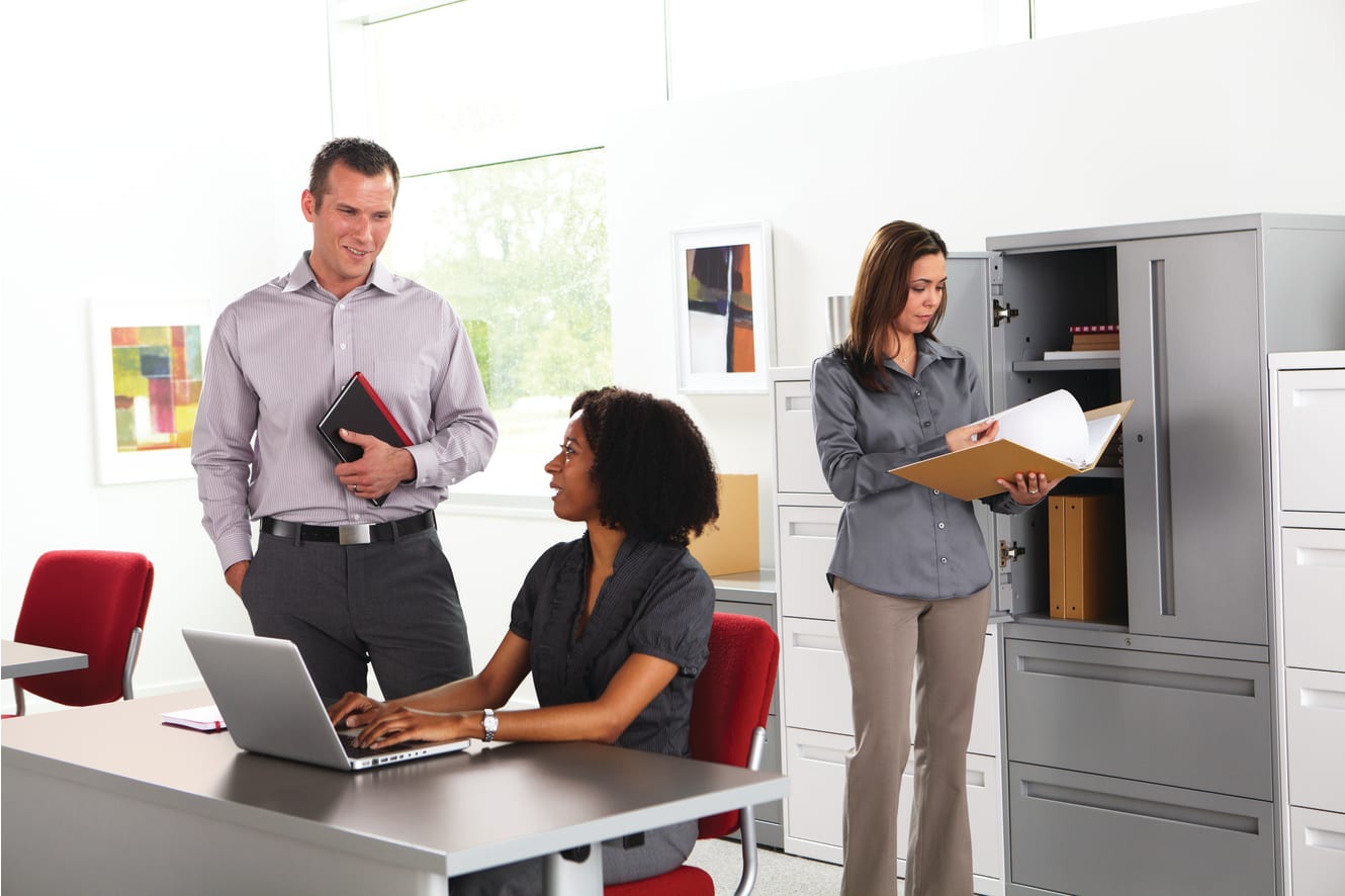 The image depicts a professional office setting with three individuals engaged in various work-related activities. In the foreground, a man and a woman are standing and conversing, while in the background, another woman is seated at a desk, working on a l