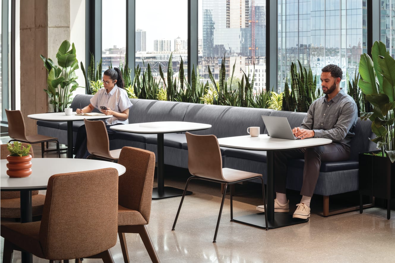Café-style seating area with upholstered banquette, round pedestal tables, and mixed-material guest chairs, complemented by integrated planter with greenery and adjacent lounge seating.