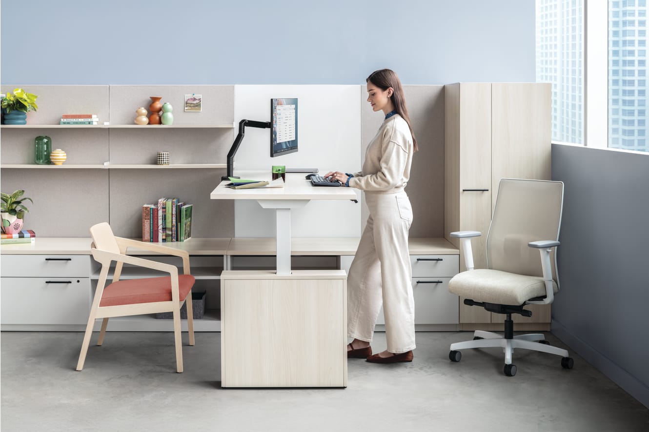 The image depicts a modern, minimalist home office setup with a woman standing at a standing desk, surrounded by shelves, chairs, and other office equipment.