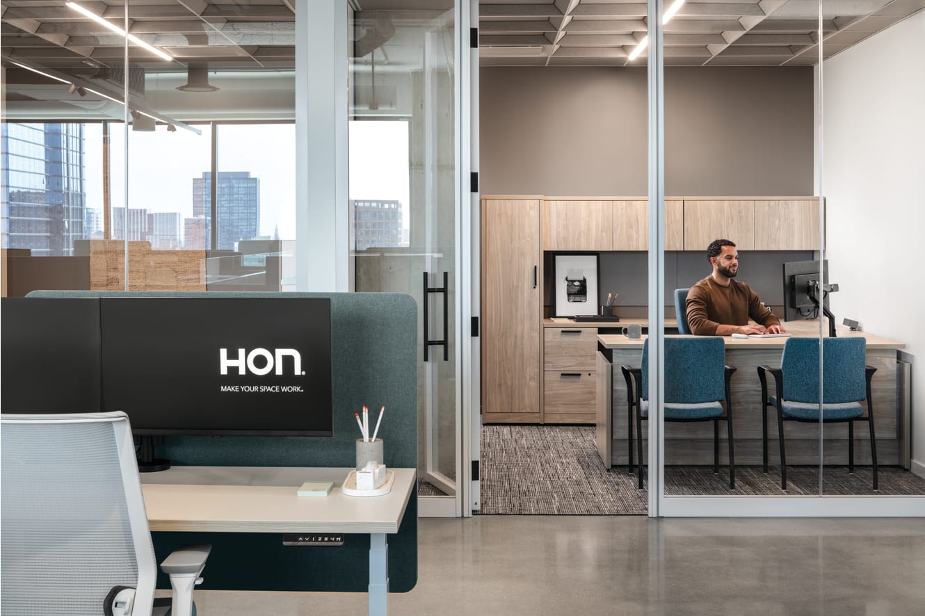 Open workstation with height-adjustable desk, privacy panel, task chair, and monitor, paired with glass-enclosed office featuring L‑shaped work surface, storage cabinets, and guest seating.