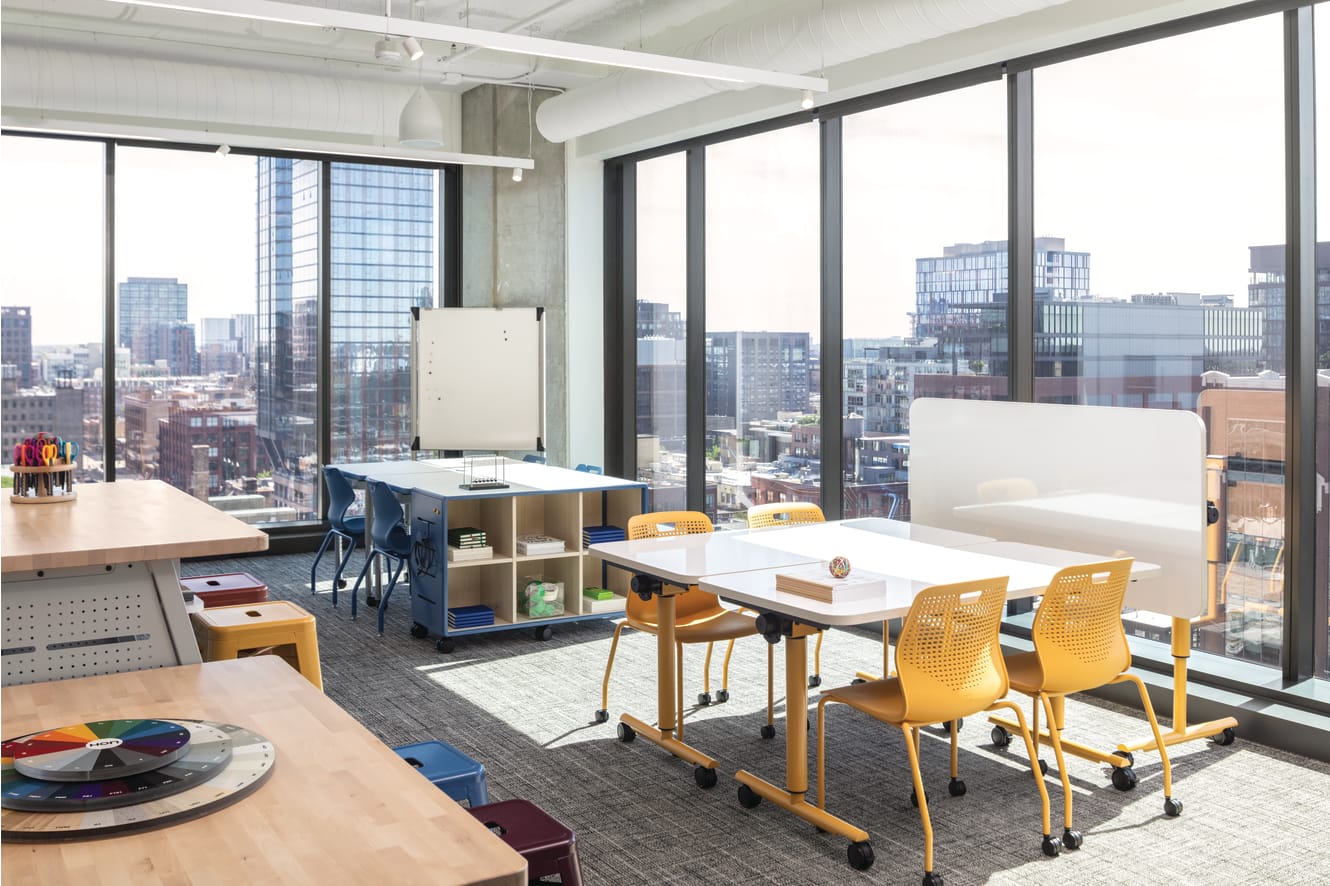 Flexible workspace with mobile tables, laminate work surfaces, caster-based storage unit, and stackable plastic chairs with perforated backs.