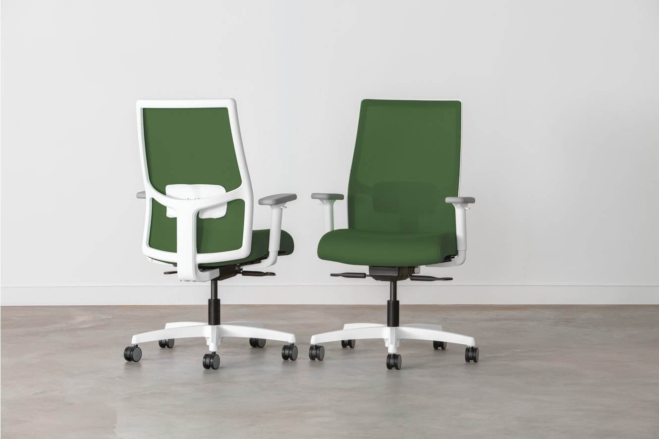 Two green and white office chairs with adjustable features are positioned in a minimalist, white-walled setting.