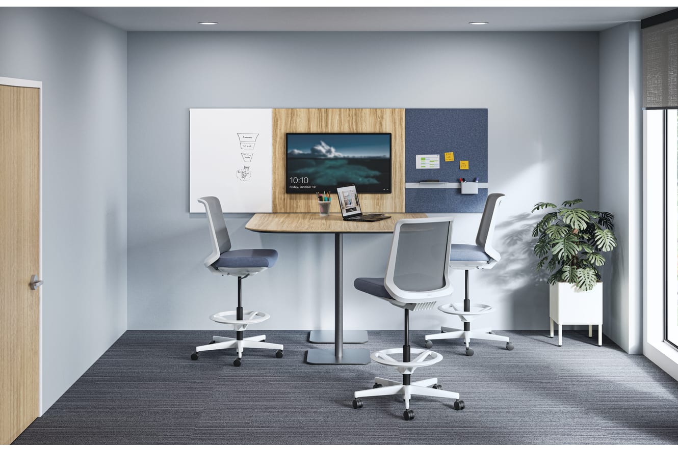 A compact collaboration room with a standing-height table, gray mesh stools, a wall-mounted display, whiteboard, and pinboard panel, accented by a large potted plant.