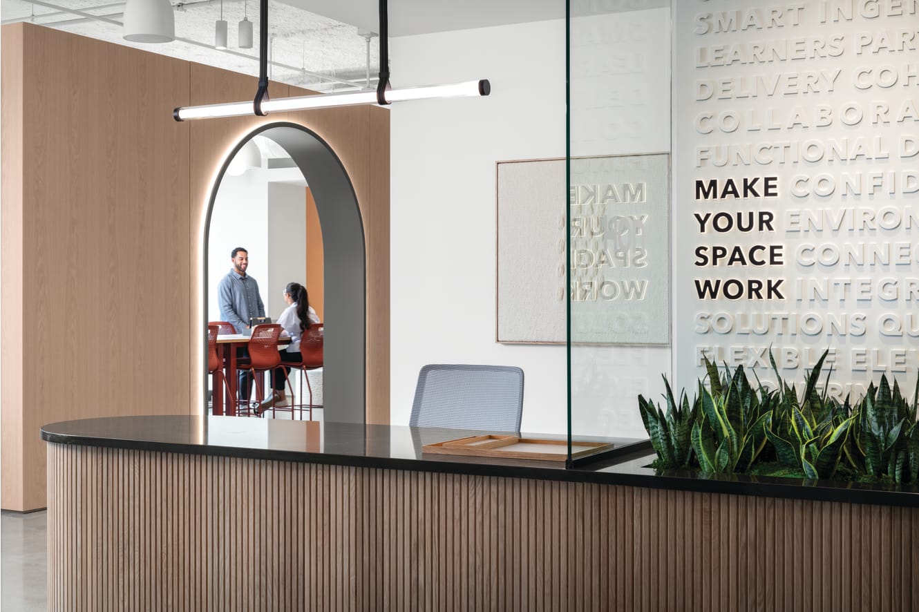 Reception counter with wood slat detailing and solid surface top, paired with task seating, integrated planter, pendant lighting, glass partition, and arched wall opening leading to collaborative seating area.