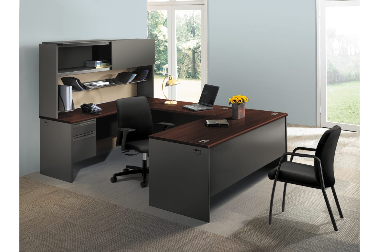A modern and minimalist home office setup with a large wooden desk, a black computer chair, and a bookshelf unit against a bright, natural-lit background.