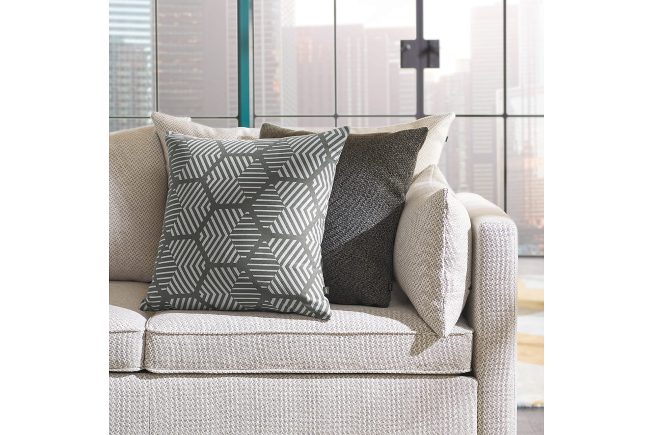 A cozy living room with a beige sofa adorned with patterned and textured throw pillows, set against a backdrop of large windows overlooking a cityscape.