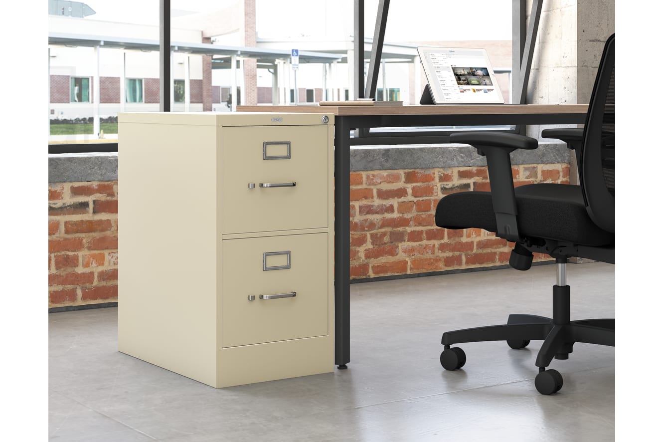 A beige filing cabinet stands in the foreground, with a desk and office chair visible in the background, set against a brick wall.