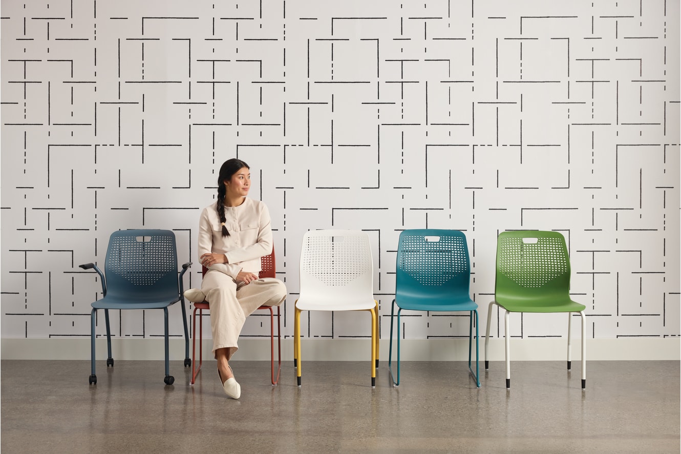 A woman in a beige outfit sits on a chair in front of a patterned wall, surrounded by colorful chairs in various shades of blue and green.