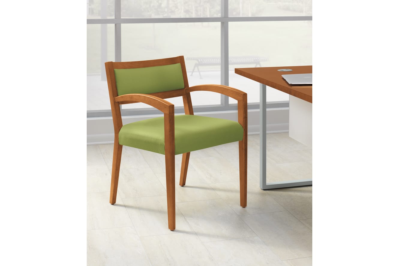 A wooden chair with a green cushioned seat sits in a bright, minimalist office environment with a large window in the background.