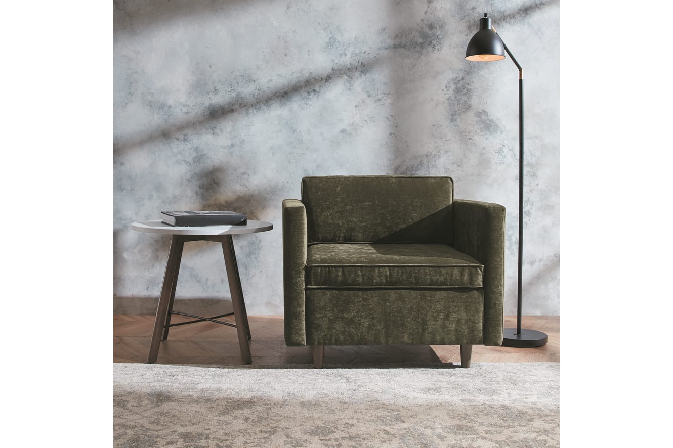 A cozy living room setting with a green upholstered armchair, a round wooden side table, and a floor lamp against a textured marble-like wall backdrop.