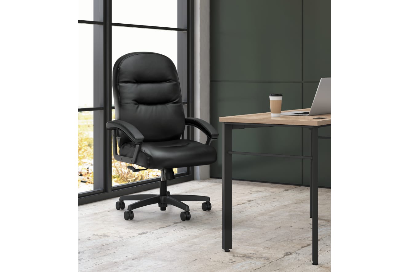 A black leather office chair sits in front of a large window, with a wooden desk and laptop visible in the background.