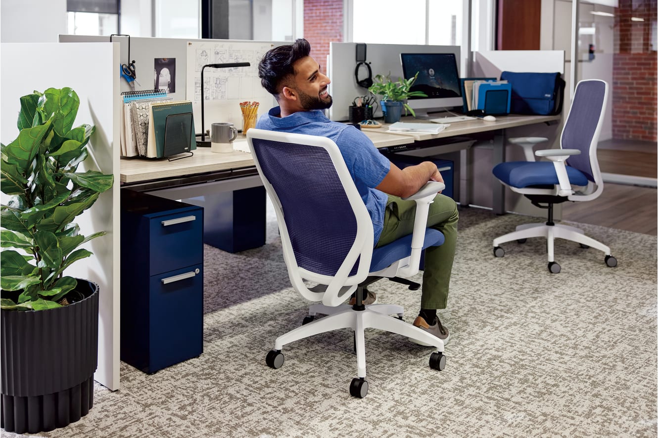 A person with a beard is sitting at a desk in an office environment, surrounded by various office equipment and a potted plant.